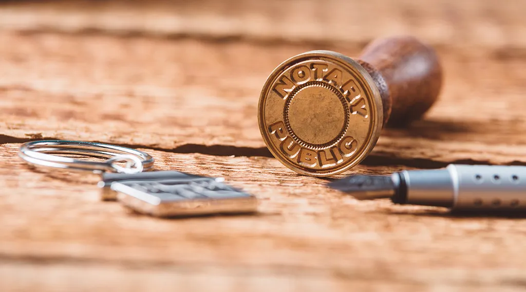 A brass Notary Public stamp lying on a wooden desk beside keys and a pen