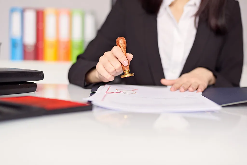 A notary in a dark jacket stamping personal documents at an office desk